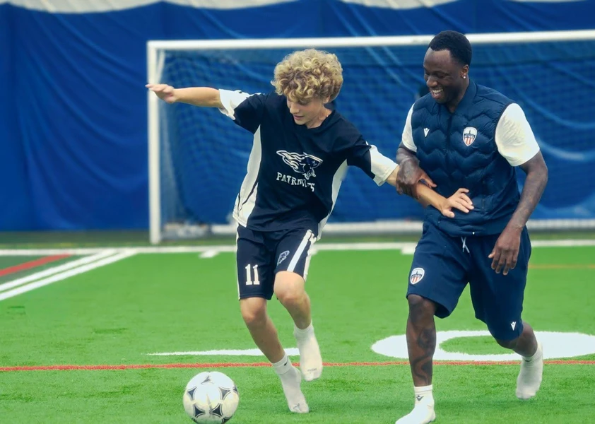 Un élève en uniforme de soccer dispute le ballon avec un entraîneur sur un terrain intérieur. Les deux participent à un exercice de jeu dans une atmosphère dynamique et conviviale, devant un filet de soccer.
